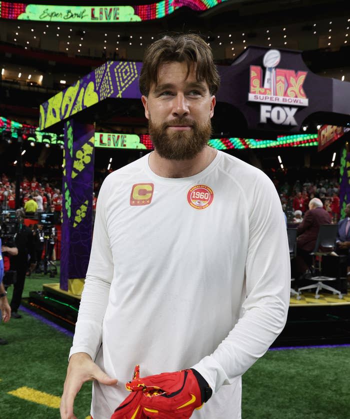 Travis Kelce in a football stadium wearing a long-sleeve shirt, holding a pair of gloves, with Super Bowl banners in the background