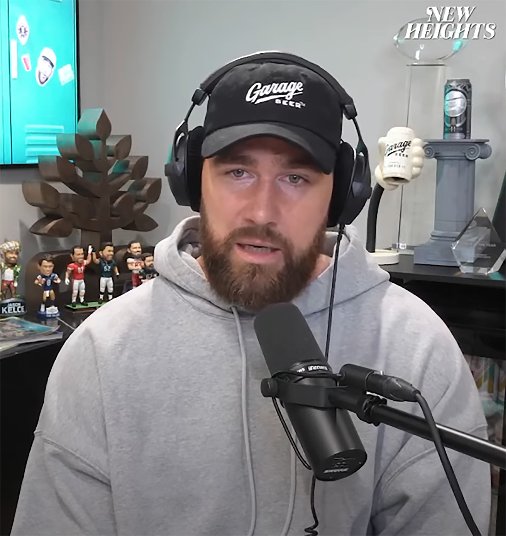 A screenshot from the New Heights podcast. Travis Kelce is talking - he is wearing a grey hoodie with a black baseball cap. His beard is long and his hair is tucked into the hat. He is sitting in front of a microphone with headphones on.