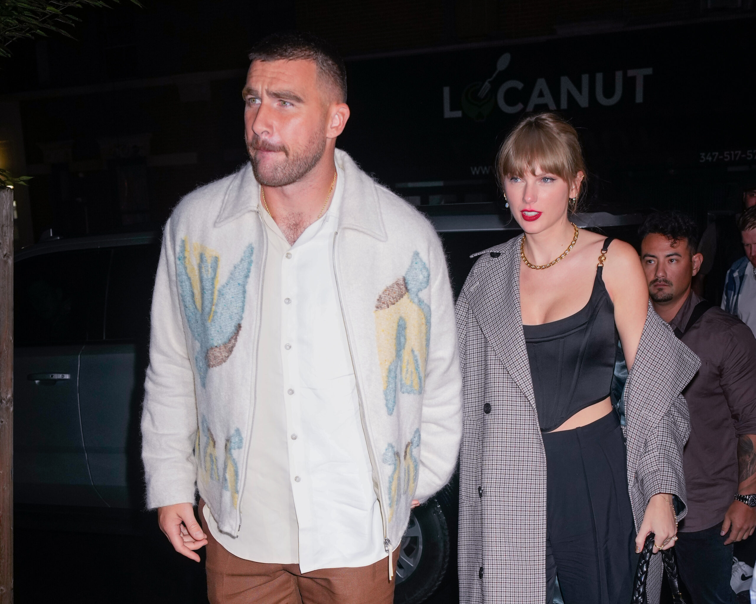 Travis Kelce with Taylor Swift. The two are walking hand in hand at night. Travis has his hair cut short, just a little longer than a buzzcut, and has a mustache. He is wearing a white button down shirt, brown slacks and a white wool jacket with soft green and yellow plant designs. Taylor is wearing a plaid overcoat hanging off one shoulder. Underneath, she has a satin black corset style top and matching black pants. She is wearing a gold chain necklace. Taylor's blonde hair is tied back in a bun.