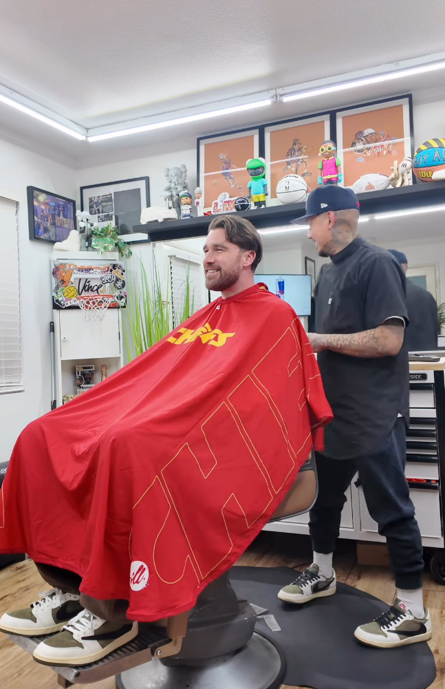 Travis Kelce sitting in a barber's chair. He has a red and yellow barber's cape draped across his shoulders. His barber is faced away from the camera - he is a bald young man covered in tattoos wearing a navy baseball cap, black tunic and pants.