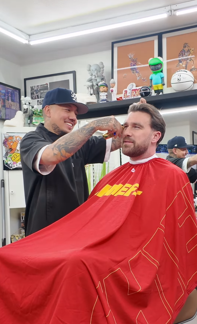 Travis Kelce getting his hair cut. His barber is trimming the sides of his head with an electric razor and smiling.