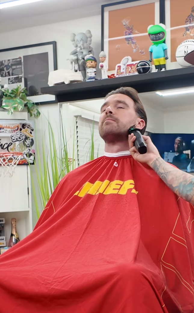 Travis Kelce getting his beard shaved down by an electric razor. His barber is not in the shot, but his tattooed arm can be seen.