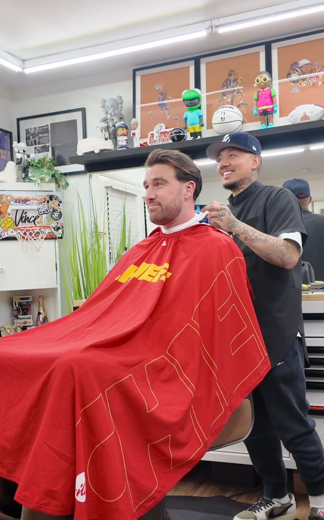 Travis Kelce sitting in a barber's chair. He has a red and yellow barber's cape draped across his shoulders. His barber is tying it around his neck.