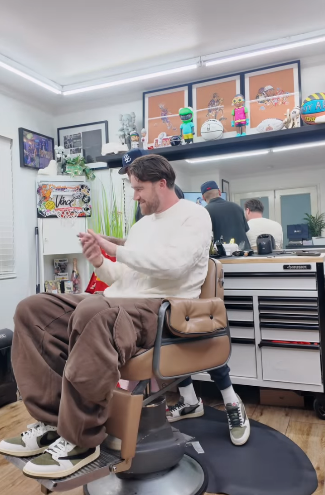 Travis Kelce sitting in his barber's chair before his haircut. He is wearing a cream long-sleeve, brown cargo pants, and cream and olive colored suede sneakers.