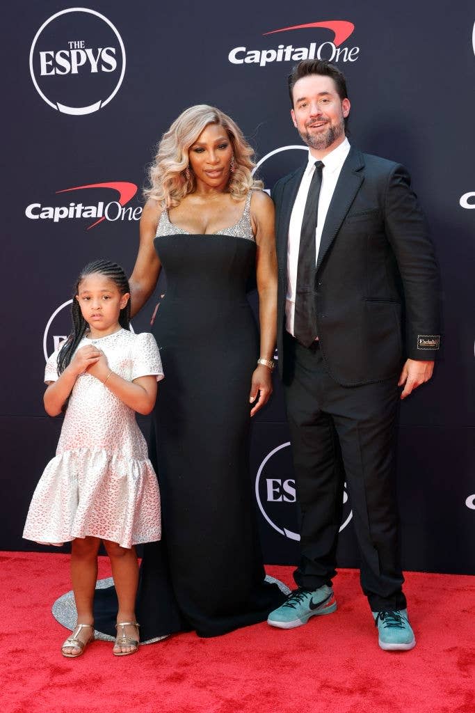 Three people on a red carpet: an adult in an elegant gown, another adult in a suit, and a child in a formal dress