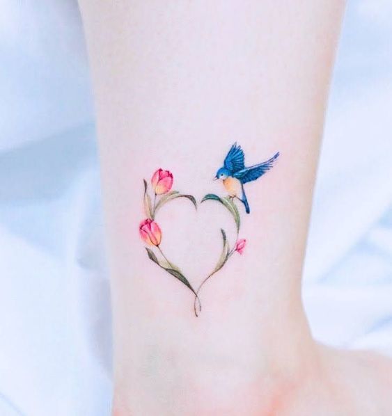 This may contain: a small tattoo on the ankle of a girl with two birds flying over flowers in the shape of a heart