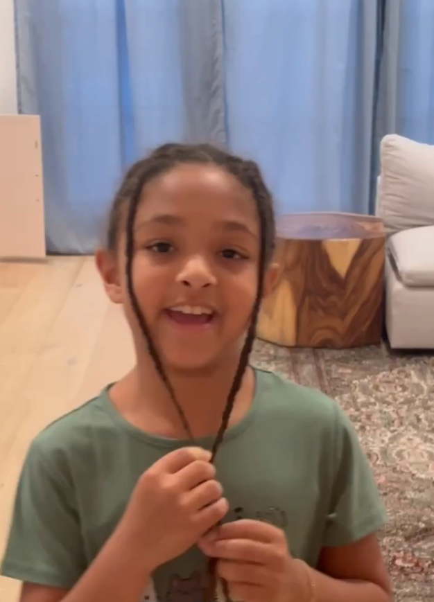 Child smiling indoors, holding braids. Text reads: "For Olympia."