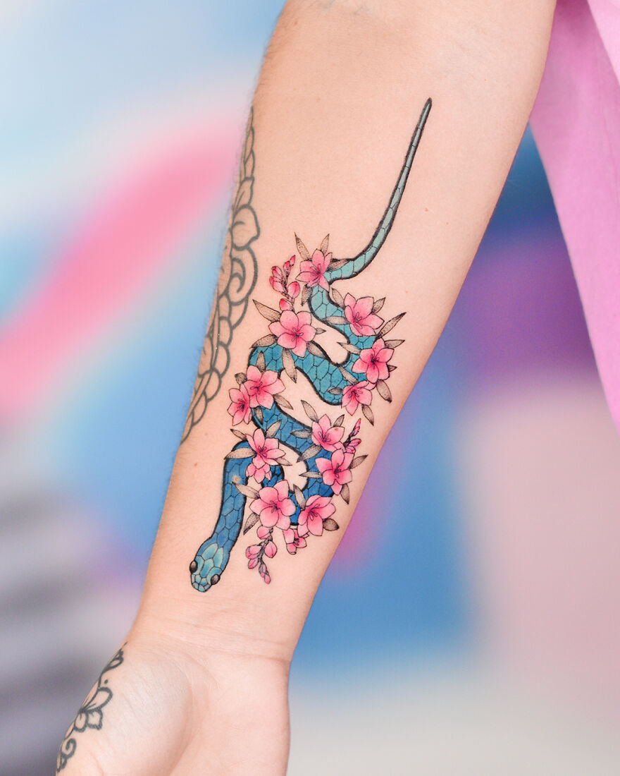 Watercolor tattoo of a blue snake intertwined with pink flowers on forearm. Watercolor tattoo of a blue snake intertwined with pink flowers on forearm.
